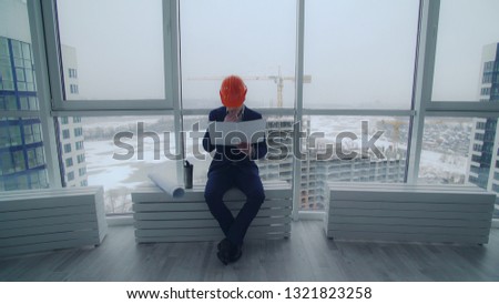 building engineer or architect have a break enjoy coffee holding documents. Handsome man wearing in elegant suit and orange hard hat. On the background construction site workers built house.
