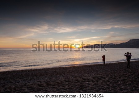 landscape of the sea and beach with people in silhouette taking pitcures on their phone of the beautiful sunset
