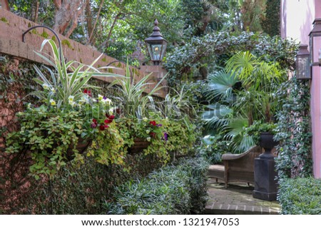 a lush brick pathway lined with greenery, flowering pots, trellised jasmine, potted palms, and a brass lantern leading to a private seating area with a wicker armchair