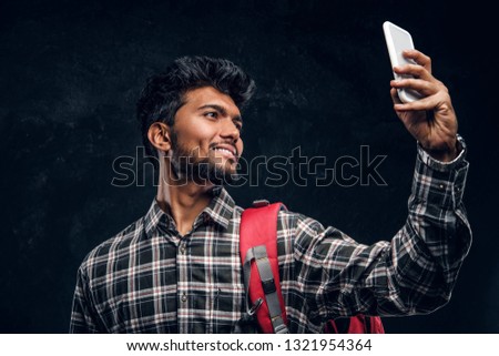 Happy Indian student with backpack taking a selfie. Studio photo against a dark textured wall