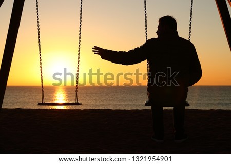 Back view backlighting silhouette of a man sitting on swing alone missing her partner at sunset on the beach in winter Royalty-Free Stock Photo #1321954901