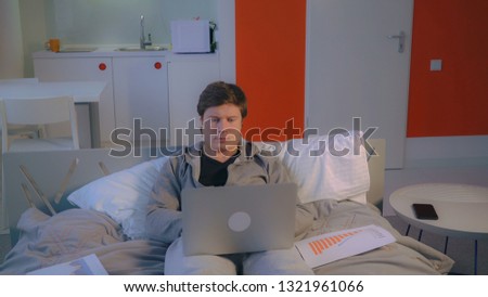 Handsome businessman working with financial report at home. Entrepreneur wearing in grey hoodie lying on bed with white pillows. Flat with modern interior and white furniture.