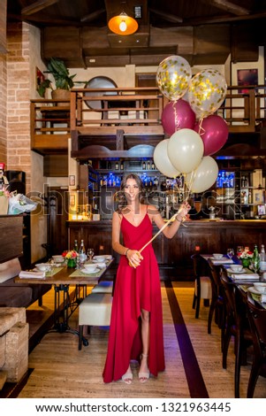 Beautiful, attractive young girl in red long dress with balloons in her hand, celebrating birthday in restaurant. Tables set in background
