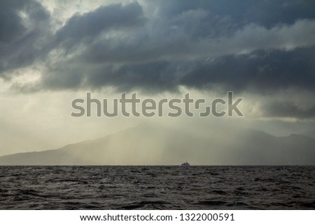Dark menacing blue clouds floating over the sea at dawn-dusk. A golden sunlight shine on a ship that is entering a rainstorm. A big mountain is faded by the rain, picturing a somewhat dramatic scene.