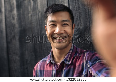 Portrait of a smiling young Asian man taking a selfie while leaning on a dark wall outside in the city