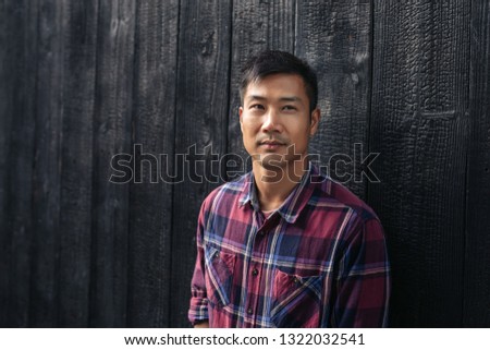 Young Asian man in a plaid shirt standing in front of a dark wall outside in the city