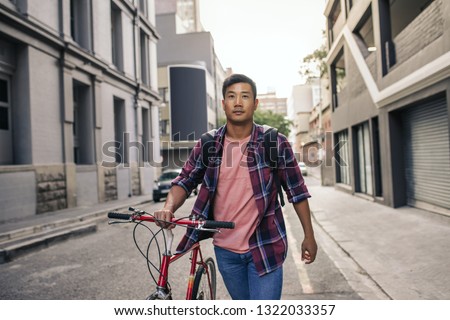 Young Asian man in a plaid shirt walking with his bicycle down an empty street in the city during his commute