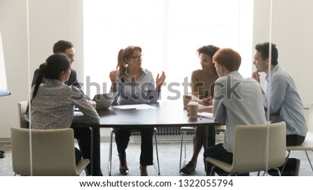 Middle aged businesswoman speaking at diverse group negotiations discussing work with colleagues, female executive talking to coworkers at corporate briefing sitting at conference table in boardroom Royalty-Free Stock Photo #1322055794