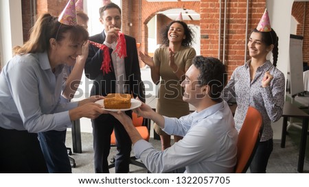 Friendly diverse employees in party hats presenting birthday cake congratulating male colleague boss, multicultural workers make surprise to coworker laughing and having fun at corporate celebration