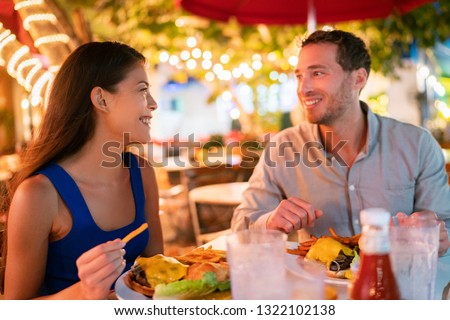 Couple eating hamburgers at outdoor restaurant terrace happy tourists on summer vacation. Florida travel people eating food at night during holidays in Miami. Asian Caucasian interracial young adults.