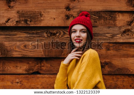 Jocund caucasian woman in funny red hat standing on wooden background. Outdoor shot of interested european lady wears cozy autumn clothes.