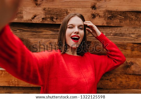 Jocund girl in red sweatshirt making selfie. Outdoor shot of joyful young female model standing on wooden background.