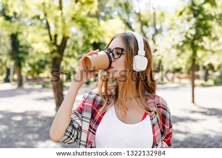 Cute lady drinking coffee with pleasure on the street. Charming female model in white headphones standing in front of trees.