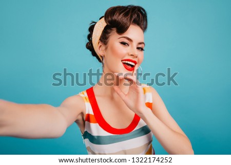 Pinup girl with bright makeup posing on blue background. Studio shot of brunette woman taking selfie.