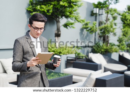 Thoughtful young asian business man working with digital tablet while holding cup of coffee in hand.Standing on a building rooftop terrace.Copy space for your text message in the right side.