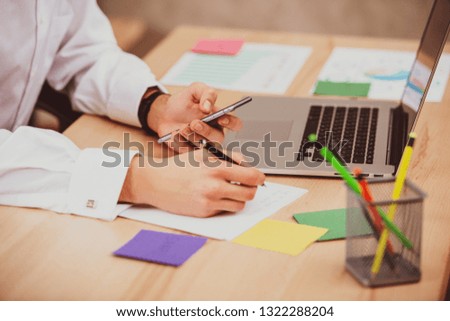 Young manager man using mobile phone in office and makes notes on paper sticker . Close-up hands, table, laptop