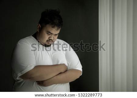 Picture of lonely obese man looks sad while standing near the window. Shot at home