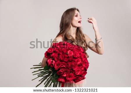 Gorgeous happy brunette with long wavy hair holding beautiful red roses bunch and eating delicious dessert with her hand. Isolated on white background in studio.