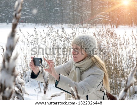 The young woman in a white jacket photographs a panorama of the coast of the winter forest lake on the phone
