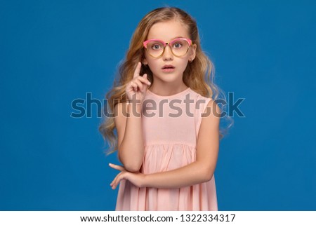 Fashionable little girl in a pink dress and glasses is standing on a blue background.