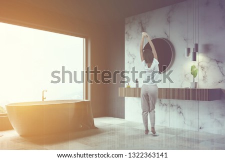 Woman in corner of luxury bathroom with gray and white marble walls, large window, white tiled floor, white bathtub and wooden sink with round mirror. Toned image doble exposure
