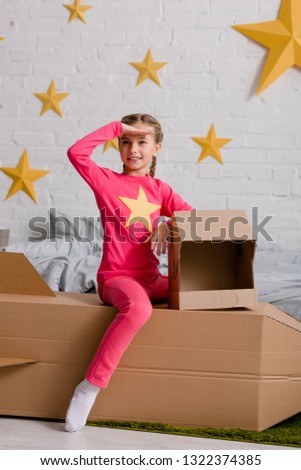 Excited kid sitting on cardboard rocket and looking away with smile