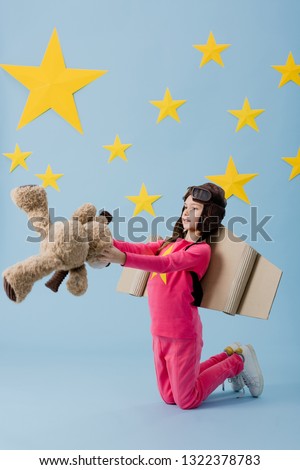 Kid with cardboard wings standing on knees and holding teddy bear on blue starry background