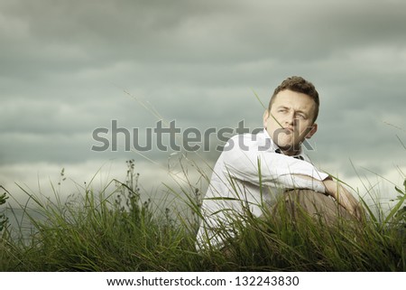 Young businessman sitting on the meadow, resting and thinking. Cloudy sky in background.