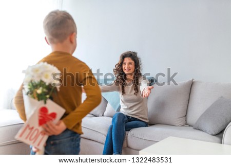 Rear view of lad with bunch of beautiful flowers behind back preparing nice surprise for his mother. Cute boy offering flowers and card to his mother in the living room 