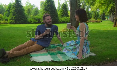 A guy with a beard with a girl in dress sitting on a park lawn on blankets on the floor and drinking coffee from plastic cups.