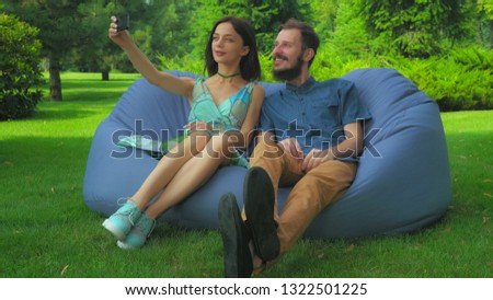 Young brunette girl and a guy with a beard sitting in a chair in the park and make selfie phone.