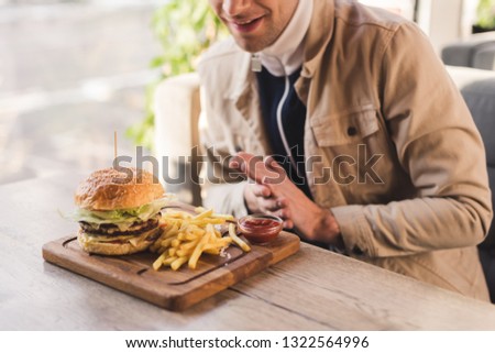 cropped view of cheerful man looking at tasty burger and french fries on cutting board in cafe