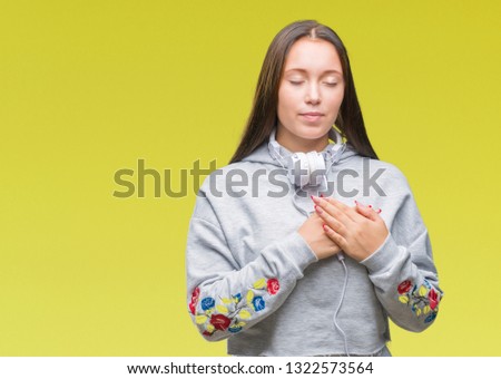 Young beautiful caucasian student woman wearing headphones over isolated background smiling with hands on chest with closed eyes and grateful gesture on face. Health concept.