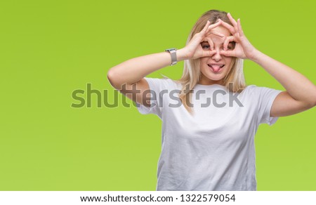Young caucasian woman over isolated background doing ok gesture like binoculars sticking tongue out, eyes looking through fingers. Crazy expression.