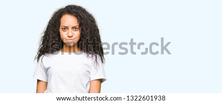 Young beautiful girl with curly hair wearing casual white t-shirt puffing cheeks with funny face. Mouth inflated with air, crazy expression.