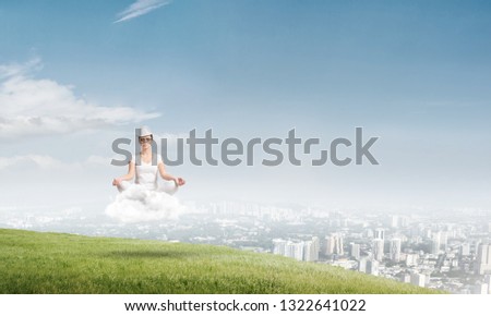 Young woman in white clothing keeping eyes closed and looking concentrated while meditating on cloud in the air with city view on background.