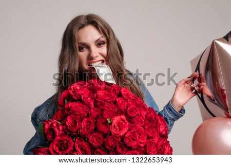 Cheeky pretty woman keeping big beautiful bouquet of red roses and balloons, biting money and looking at camera. Crazy young model getting presents on birthday and posing on isolated background.