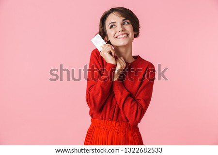 Portrait of a beautiful young woman wearing red clothes standing isolated over pink background, holding credit card