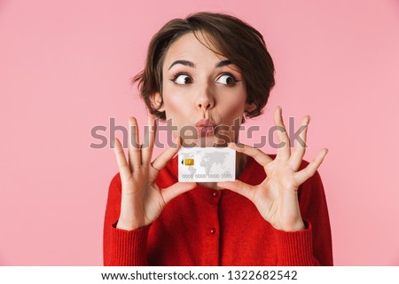 Portrait of a beautiful young woman wearing red clothes standing isolated over pink background, holding credit card Royalty-Free Stock Photo #1322682542