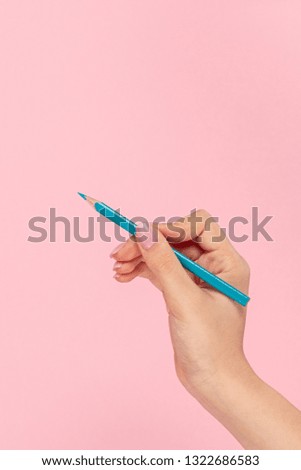 Crop female hand holding coloring pencil of blue color and writing on pink background 