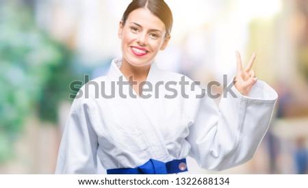 Young beautiful woman wearing karate kimono uniform over isolated background smiling with happy face winking at the camera doing victory sign. Number two.