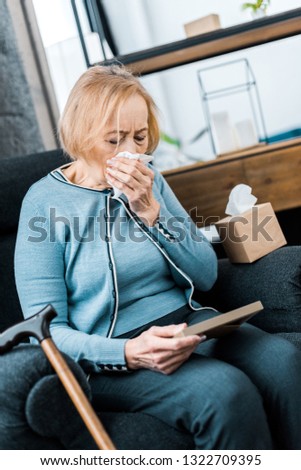sad senior woman crying and wiping face from tears with tissue while looking at picture frame 