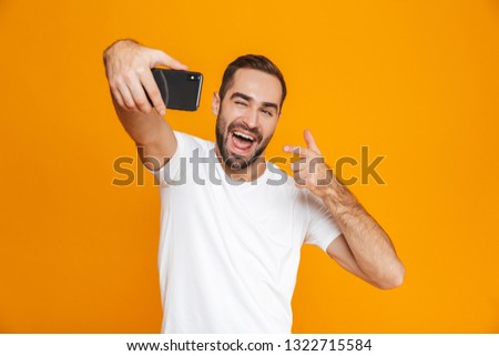 Photo of satisfied guy 30s in casual wear laughing and taking selfie on cell phone isolated over yellow background