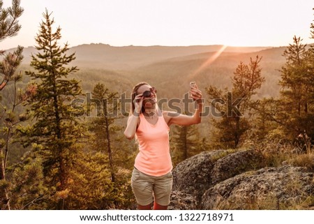 thirty-year-old smiling woman standing in the mountains and takes a selfie photo on your smartphone. A selfie at sunset in the summer