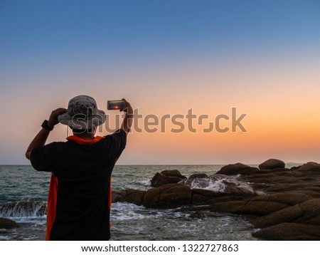 Man taking photos of sunset with mobile phone. Traveler young man is shooting video of beautiful sunset landscape on mobile phone camera. Summer vacation travel and holiday.