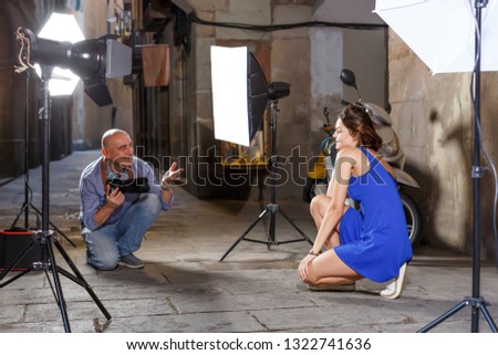 Photographer using professional camera and light equipment for taking pictures of brunette woman in blue dress on town street