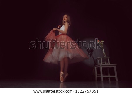 Young beautiful fair-haired white blond ballerina in a pink ballet skirt dances against a black wall and a bouquet of flowers illuminated by a light source
