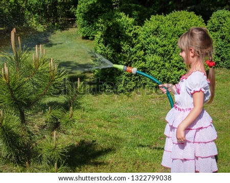 Pretty little girl 5 year old with long blond hair in lovelly white dress watering a small pine tree in the garden. Beautiful green background of spring garden. Ecology concept