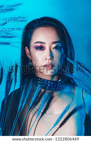 Young tender asian woman with artistic make-up and wheat in hair looking at camera isolated on blue background