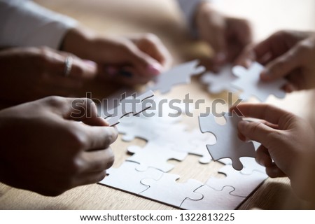 Close up members of multiracial team assembling white puzzle, African American and Caucasian people searching solution together, team building activity, staff training, support and unity Royalty-Free Stock Photo #1322813225